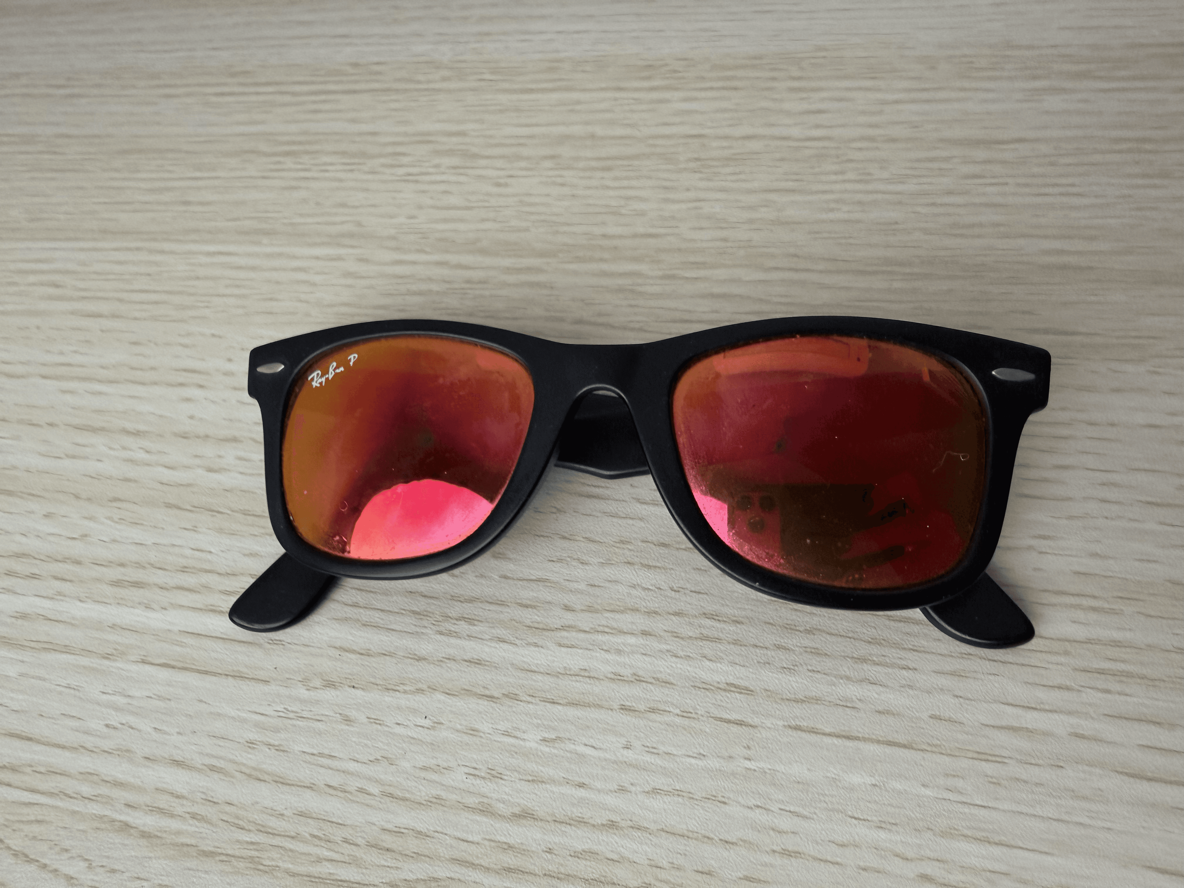 Chris’s black Ray-Ban frames with red-orange mirrored lenses resting on a pale wooden surface.
